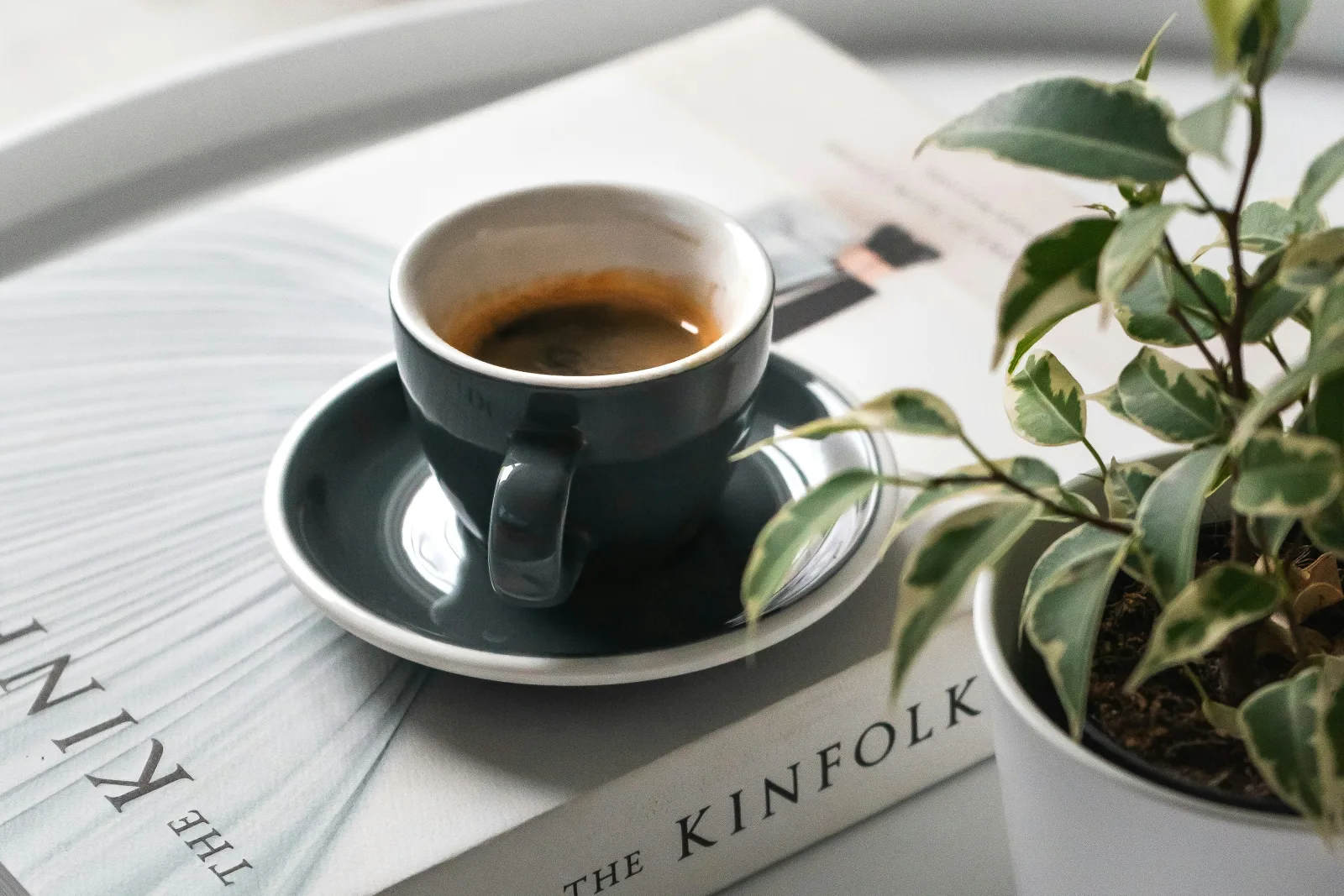 Xícara de café escura ao lado de um livro e uma planta em vaso sobre superfície branca, sugerindo reflexão sobre gestão de clínicas.
