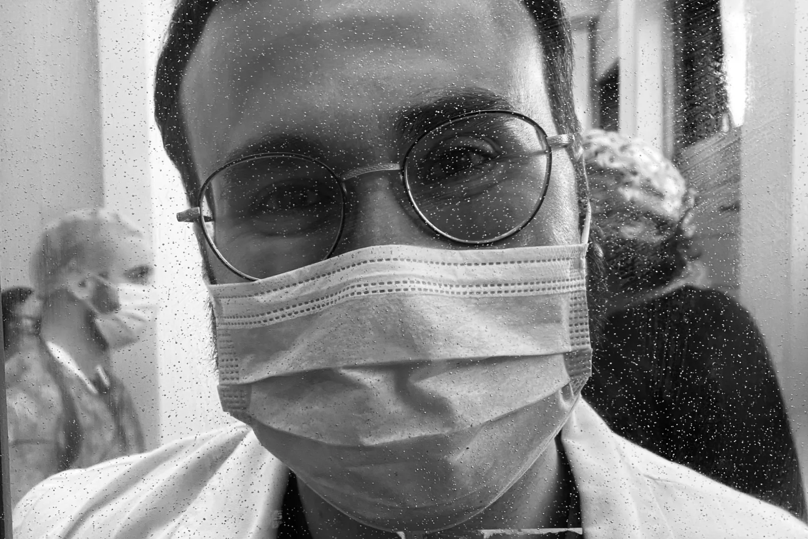Profissional de saúde de máscara e óculos, visto através de superfície molhada. Simboliza a pressão e a reflexão interna do médico.