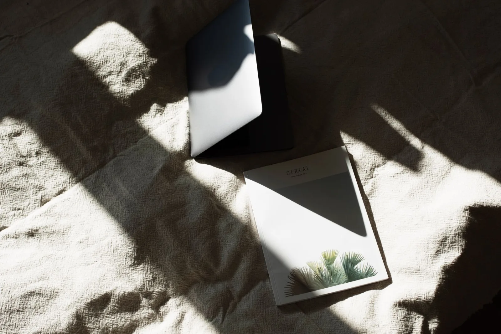 Laptop parcialmente aberto e revista sobre superfície clara, com luz e sombra natural. Ideal para reflexão e preparo.