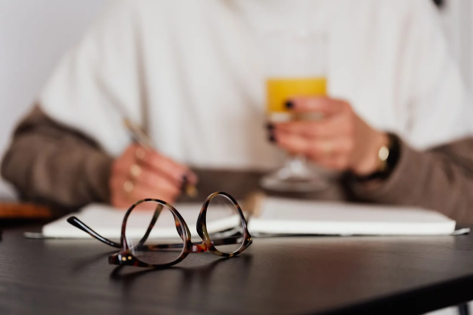 Pessoa escrevendo em caderno com óculos e suco, simbolizando planejamento e organização de processos em clínicas. Detalhe de mesa escura.
