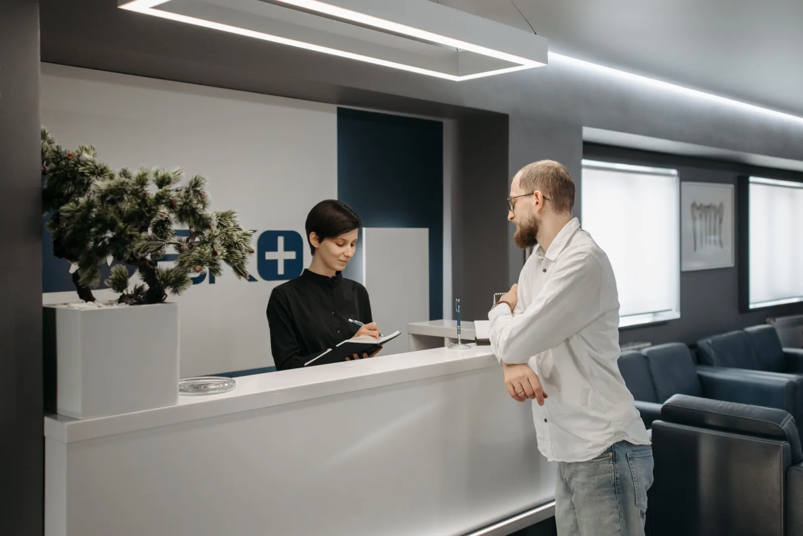 Recepcionista de clínica anota informações enquanto paciente se inclina no balcão, representando o atendimento e a escuta ativa.