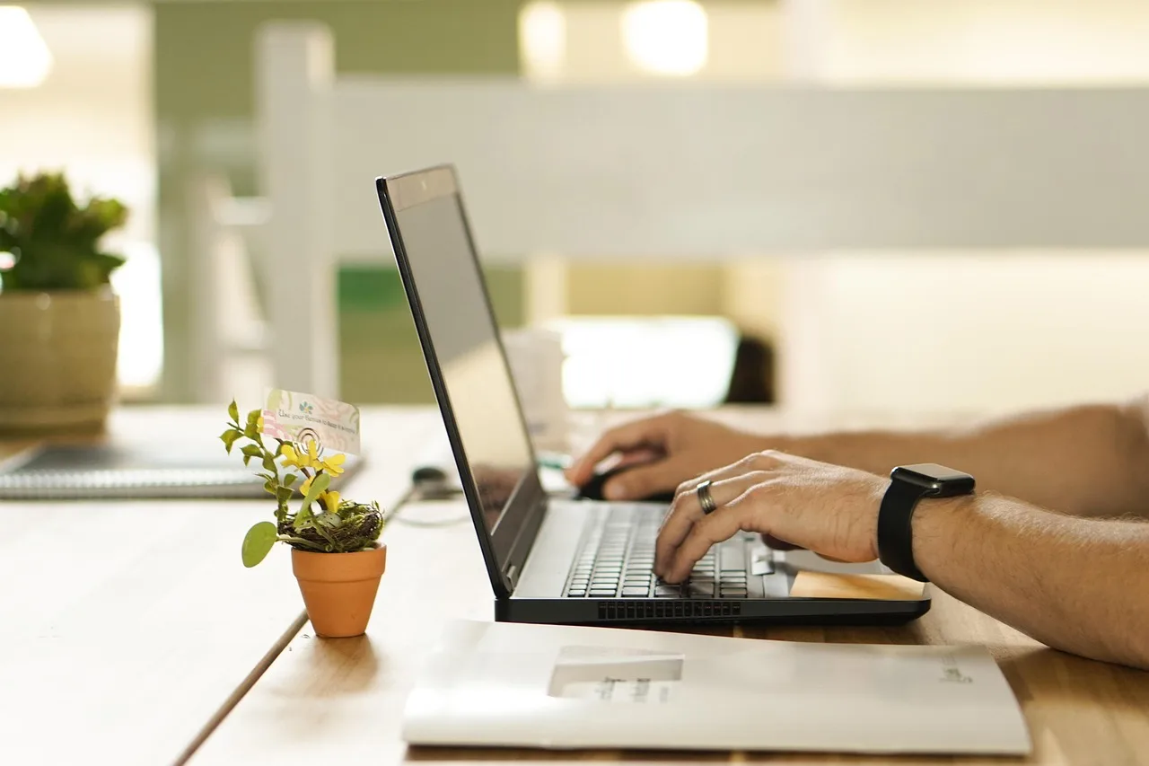 Mãos masculinas digitando em um laptop prateado sobre uma mesa de madeira clara, com documentos e um pequeno vaso de planta ao lado, simbolizando a gestão de dados e conformidade LGPD em clínicas.