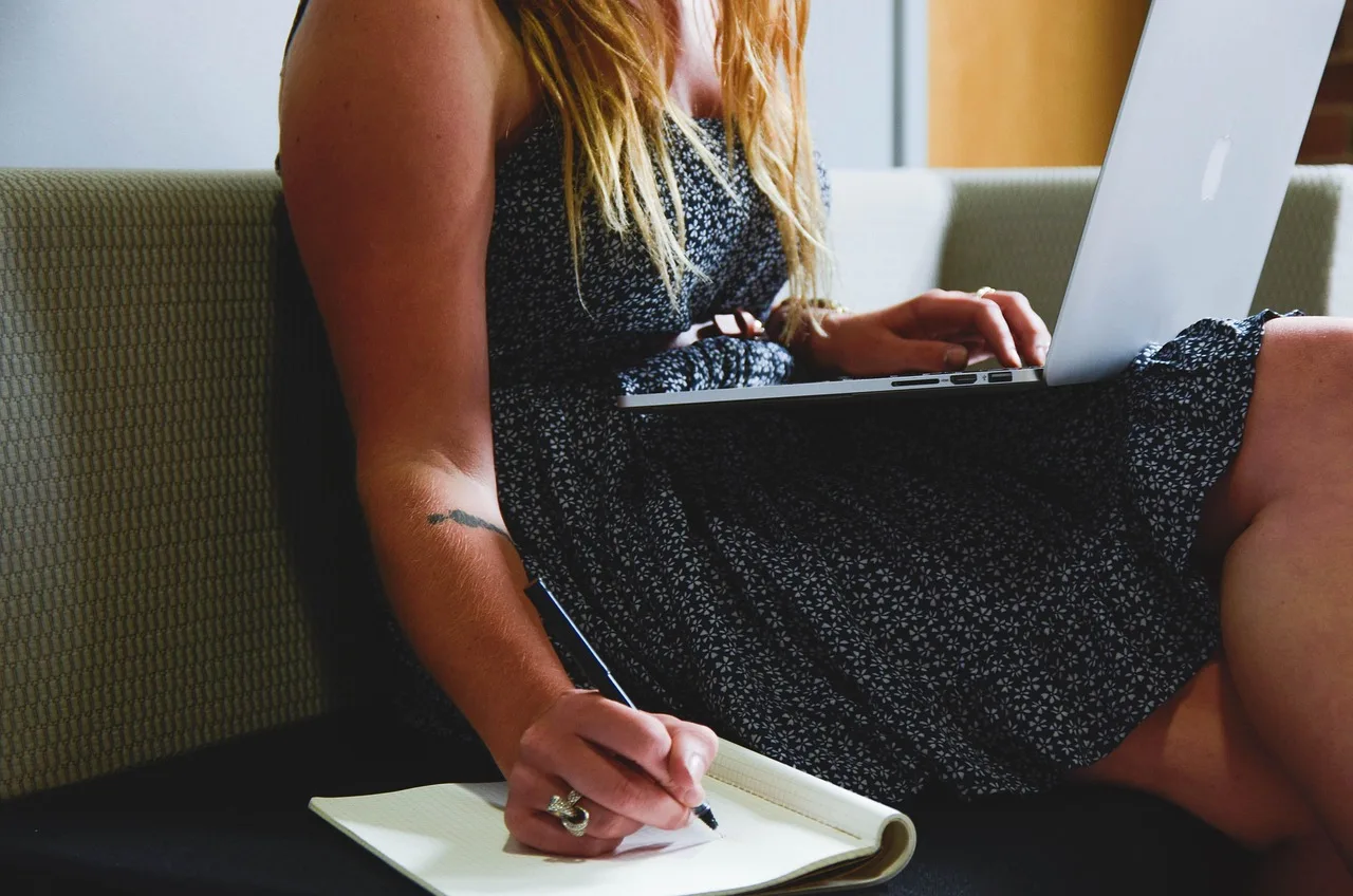 Mulher loira em vestido estampado trabalha em laptop e anota em caderno, sentada no sofá, avaliando software para gestão de clínicas.