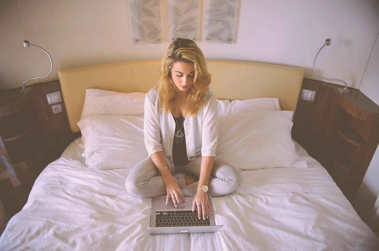 Mulher loira sentada de pernas cruzadas em uma cama branca, digitando em um laptop, simbolizando a gestão eficiente de horários por IA.