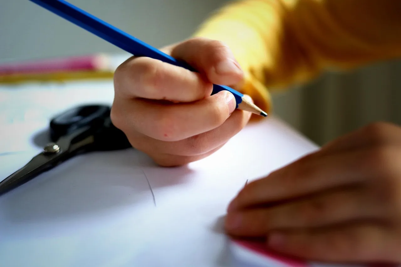 Uma mão segura um lápis azul, escrevendo em uma folha de papel branca, simbolizando a documentação manual de processos em clínicas de saúde.