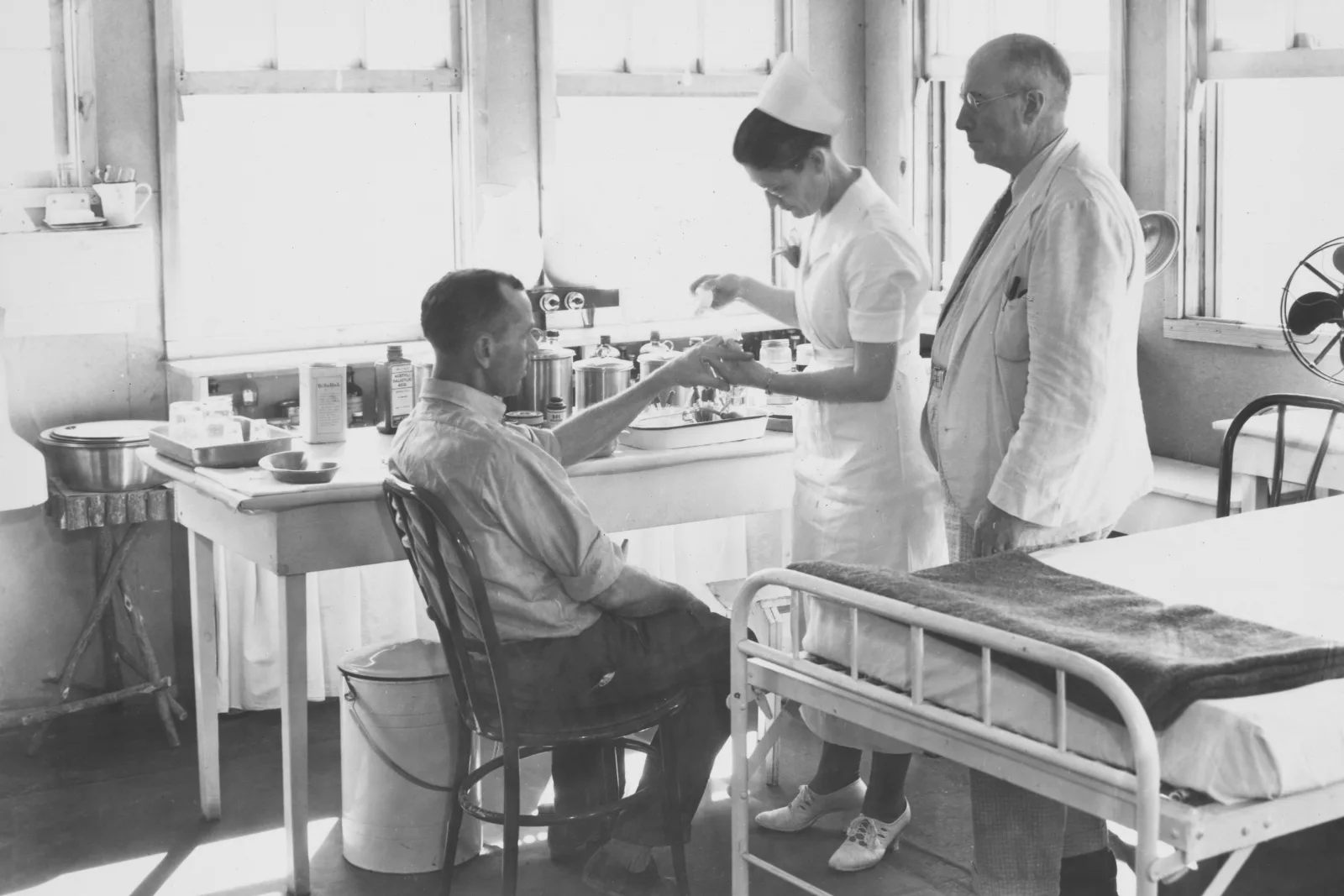 Enfermeira atende paciente sentado enquanto médico observa, ilustrando o trabalho em equipe essencial para clínicas.