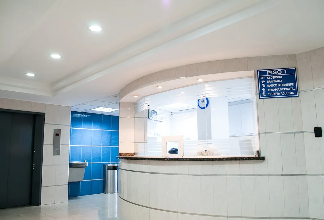 Recepção de clínica médica moderna com balcão curvo, elevador e azulejos azuis, ilustrando o ambiente de saída do paciente.