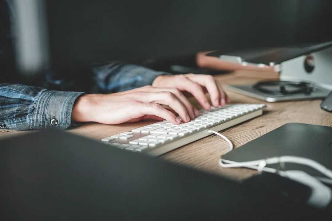 Mãos digitando em um teclado branco sobre uma mesa de madeira escura, representando a interação humana com sistemas digitais e a precisão necessária.